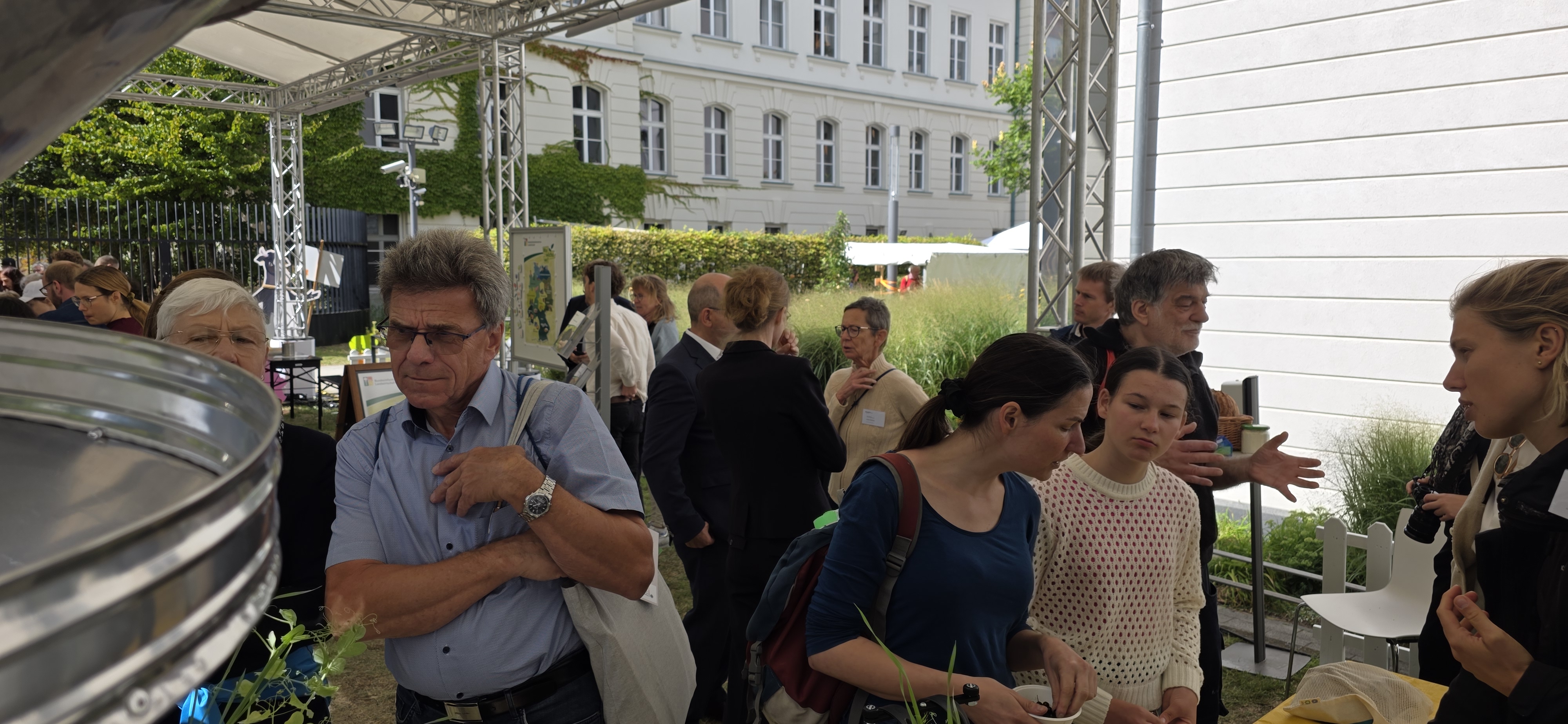 Content_Bild_2 in einem Garten im Hof - Links begutachten Menschen ein metallenes Gefäß, rechts und hinten mehrere Menschen im Gespräch vertieft
